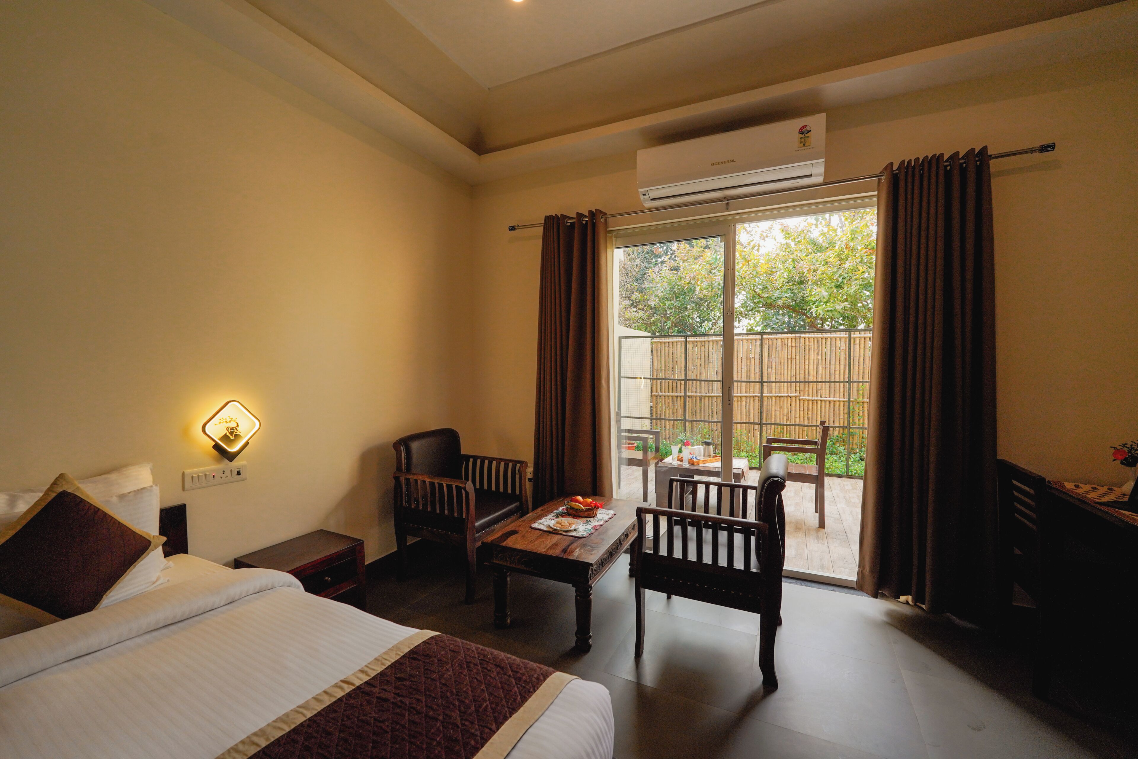 A hotel room with a bed, a desk, two chairs, a table with fruit, and a sliding glass door to a balcony.