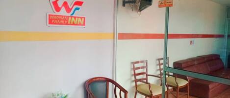 Ein Empfangsbereich mit dem Logo von 'Warisan Family Inn', einer Glastrennwand, einer Couch und einem kleinen Tisch mit einer Pflanze.
