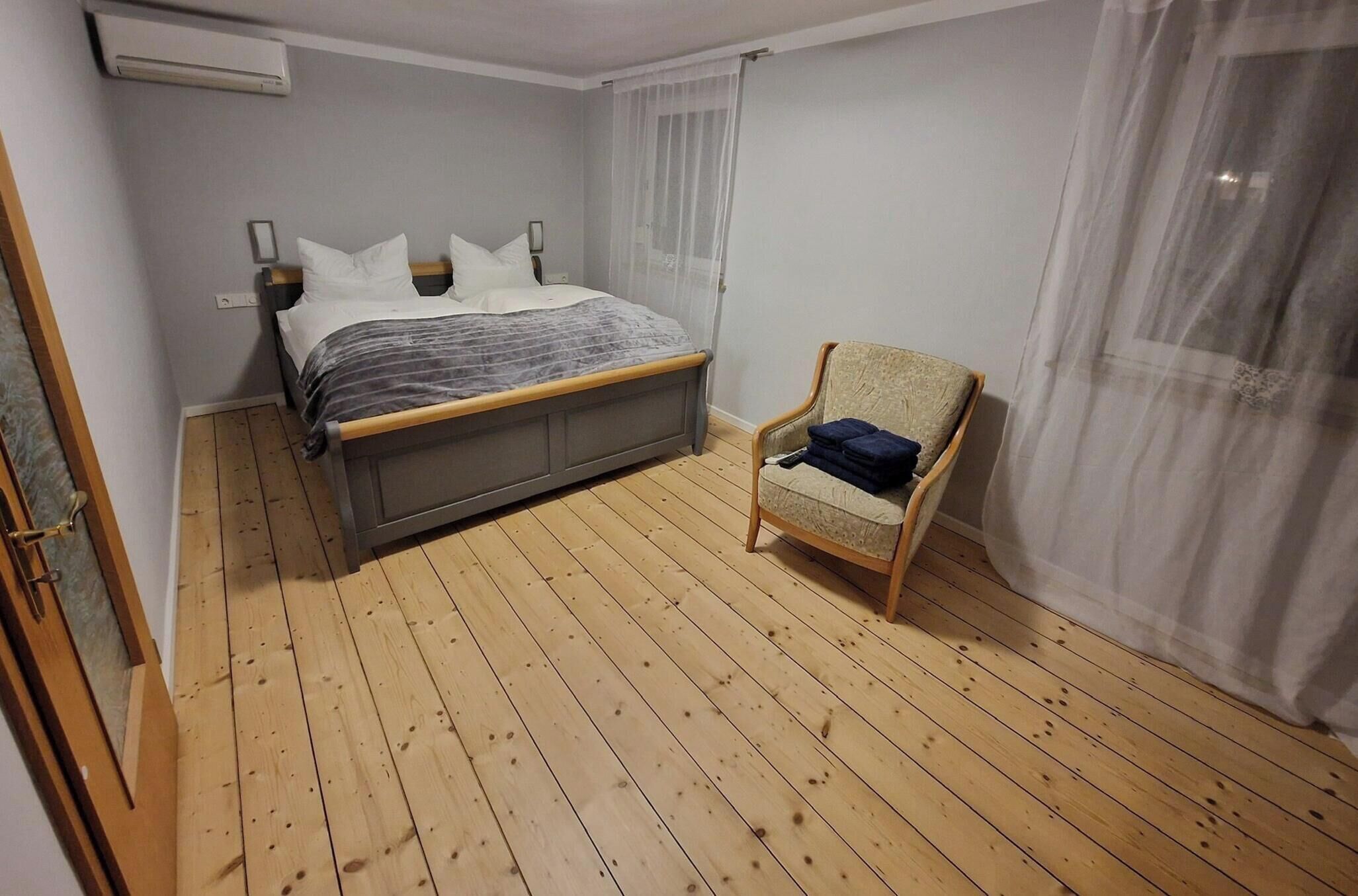 Un dormitorio con piso de madera, una cama con cabecera gris, una silla y una ventana con cortinas translúcidas.