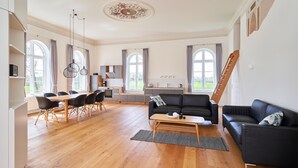 Amplia sala de estar con piso de madera, un sofá de cuero negro, una mesa de comedor con sillas y ventanas grandes con cortinas.