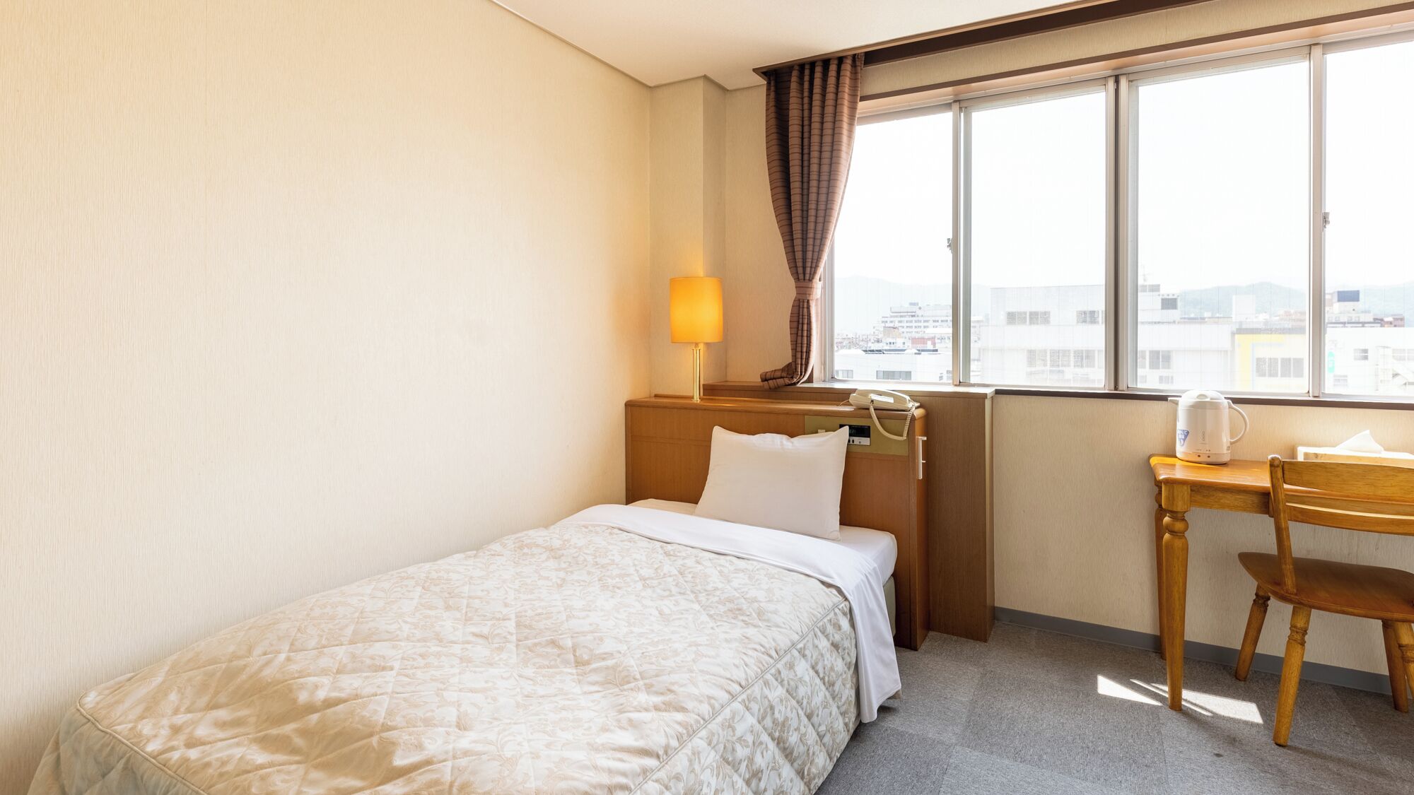 Une chambre d’hôtel avec un lit simple, un bureau avec une chaise, une fen être donnant sur des immeubles et une lampe sur le bureau.