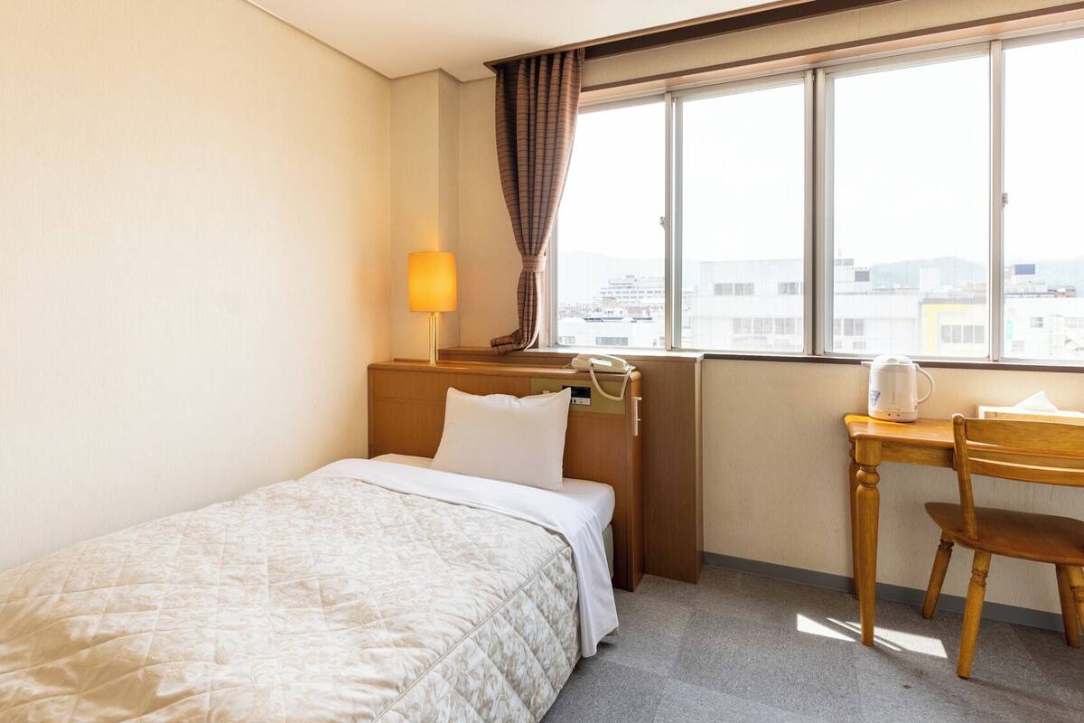Habitación de hotel con una cama individual, un escritorio con silla, una ventana con vistas a edificios y una lámpara sobre el escritorio.