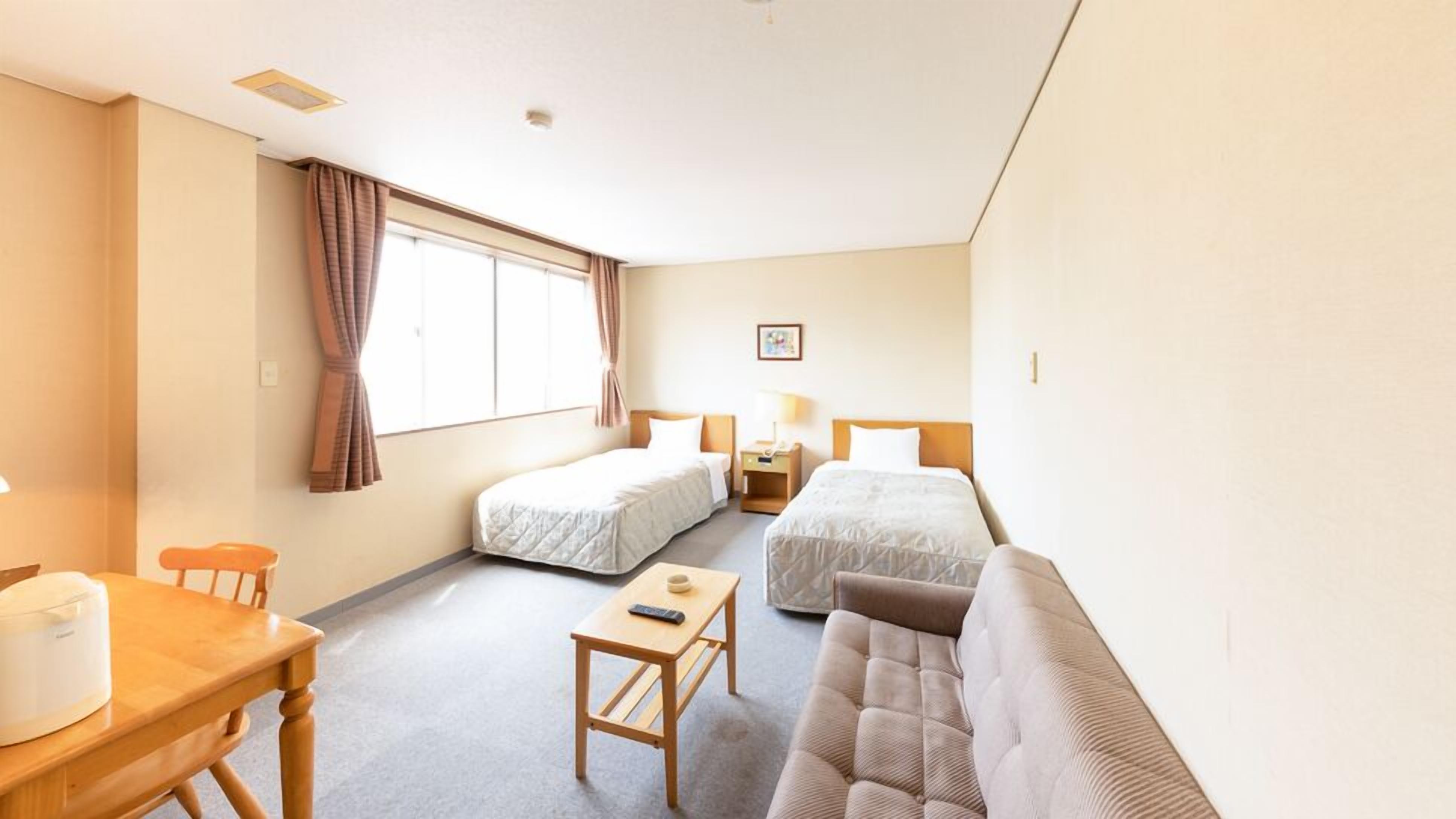 A hotel room with two single beds, a sofa, a small table, a lamp, and a window with curtains.