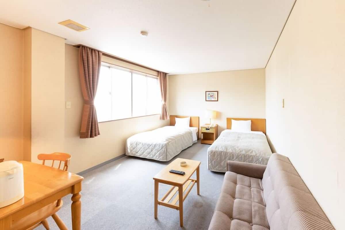 A hotel room with two single beds, a sofa, a small table, a lamp, and a window with curtains.