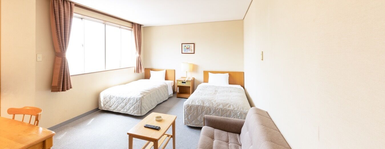 Une chambre d’hôtel avec deux lits simples, un canapé, une petite table avec une télécommande, une table de chevet avec une lampe et une fenêtre avec des rideaux.