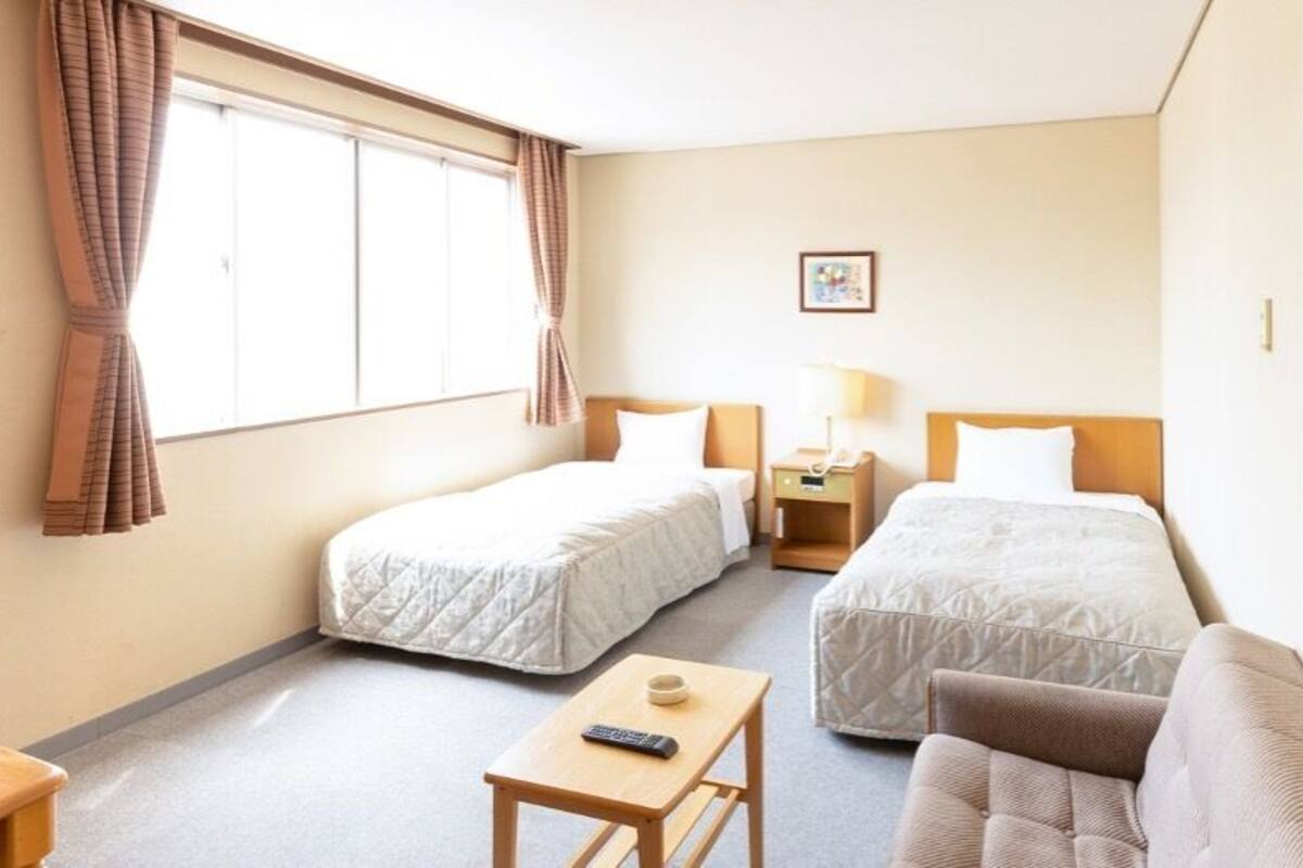 Habitación de hotel con dos camas individuales, un sofá, una mesita con un control remoto, una mesita de noche con lámpara y una ventana con cortinas.