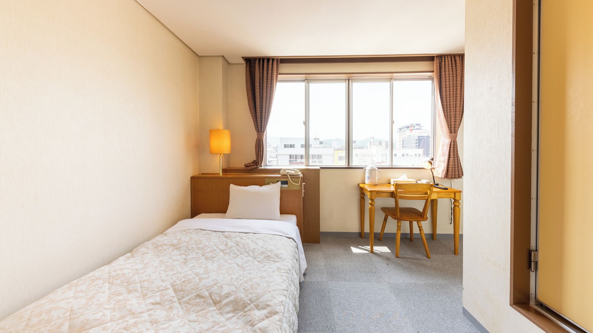 Une chambre d’hôtel avec un lit, un bureau, une chaise, une lampe et une fenêtre avec des rideaux.