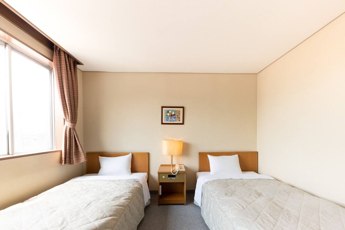 Habitación de hotel con dos camas, una mesita de noche con lámpara, una pequeña mesa y un cuadro en la pared.