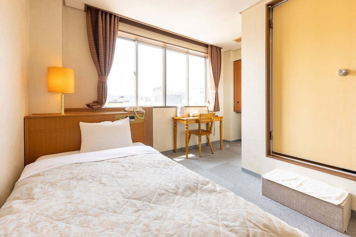 Una habitación de hotel con cama, escritorio, silla, lámpara y ventana.