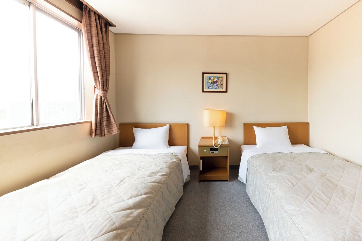 A hotel room with two beds, a nightstand with a lamp, a painting on the wall, and a window with curtains.
