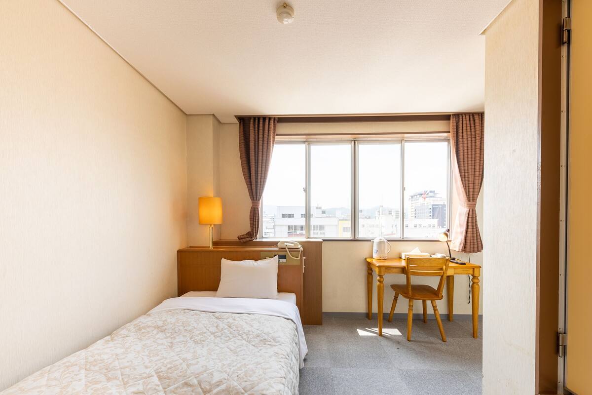 Una habitación de hotel con cama, escritorio, silla, lámpara y ventana con cortinas.