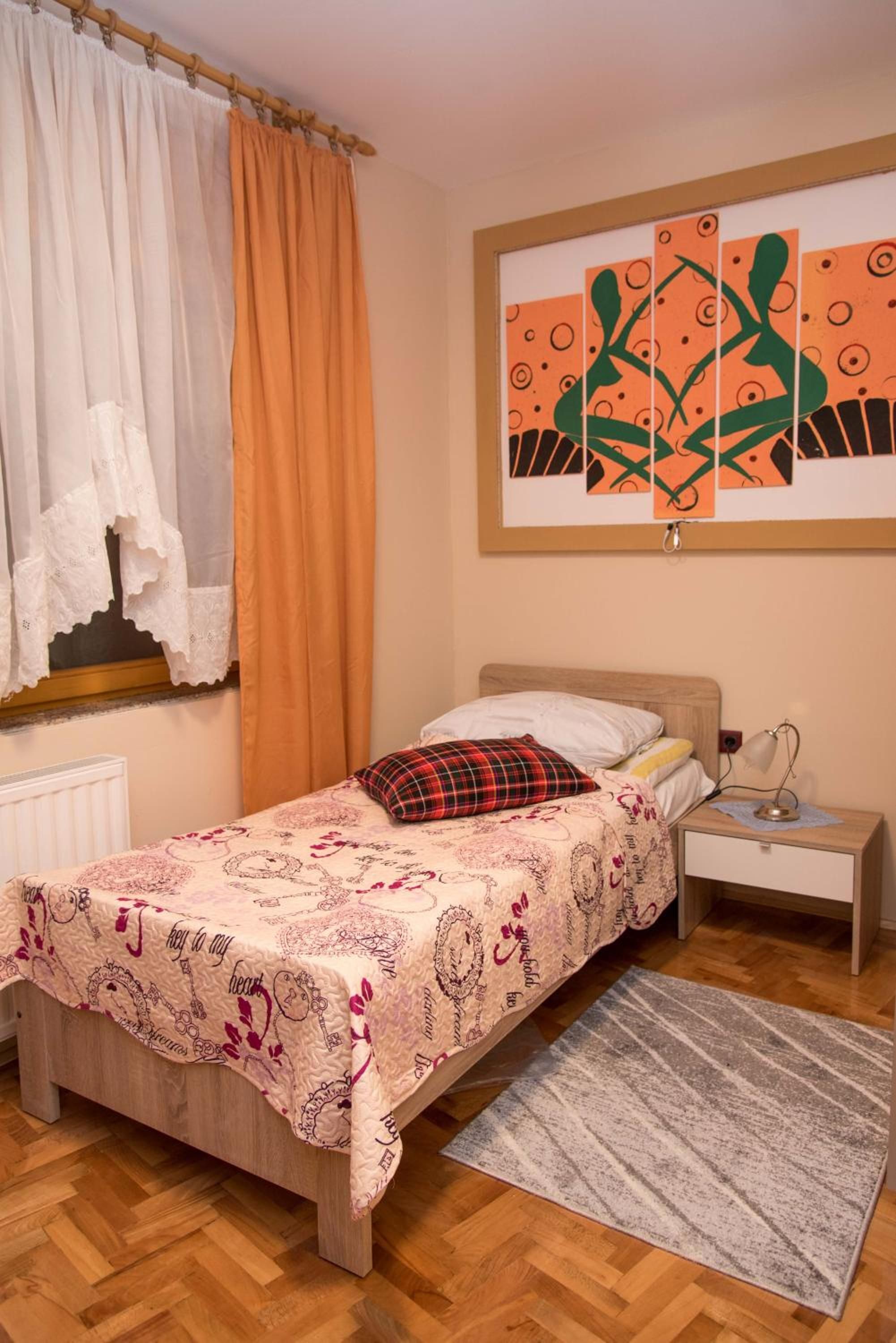 A bedroom with a bed, a nightstand, a lamp, a window with curtains, and a wall with artwork.