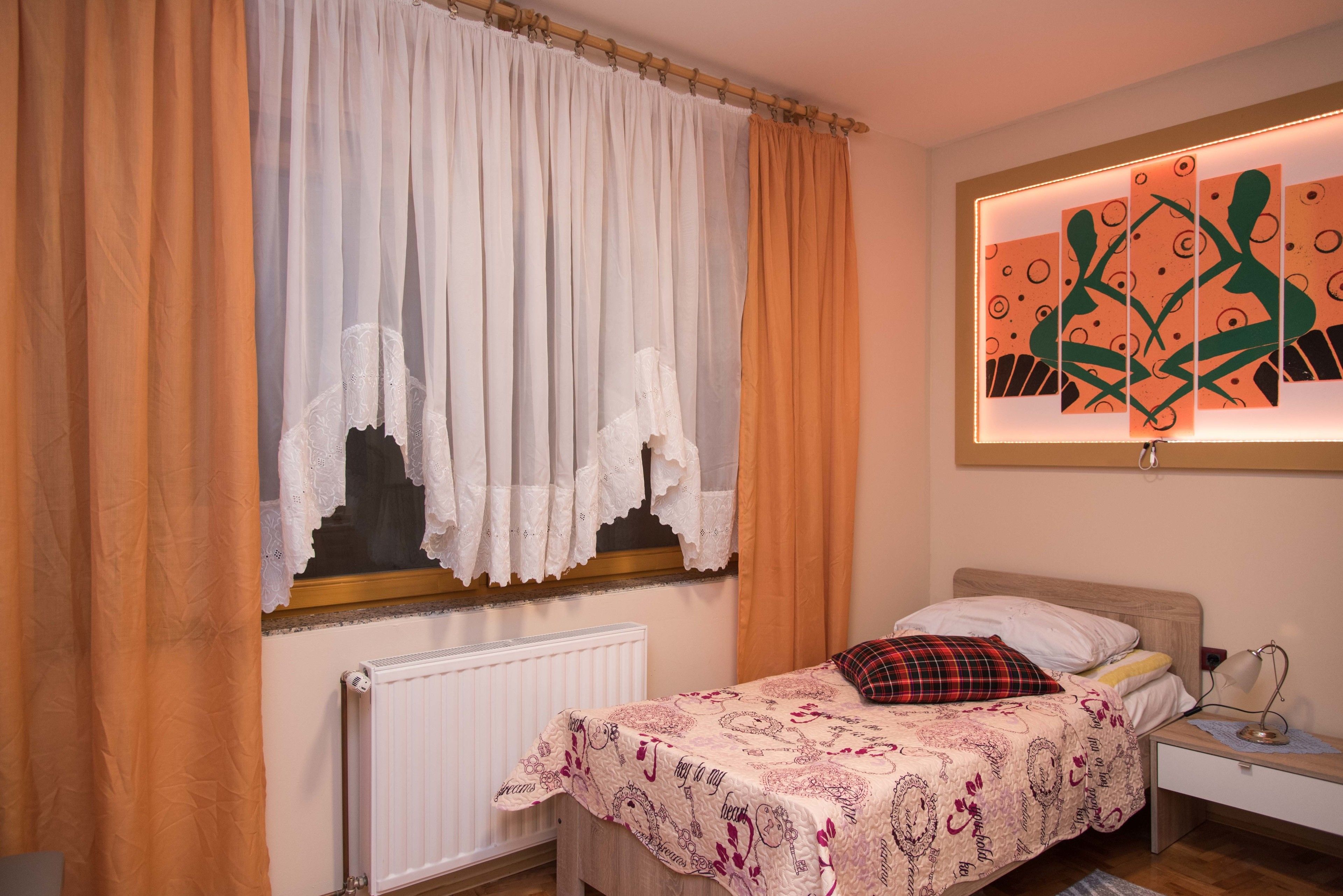 Un dormitorio con cama, mesita de noche, lámpara, ventana con cortinas y una obra de arte en la pared.