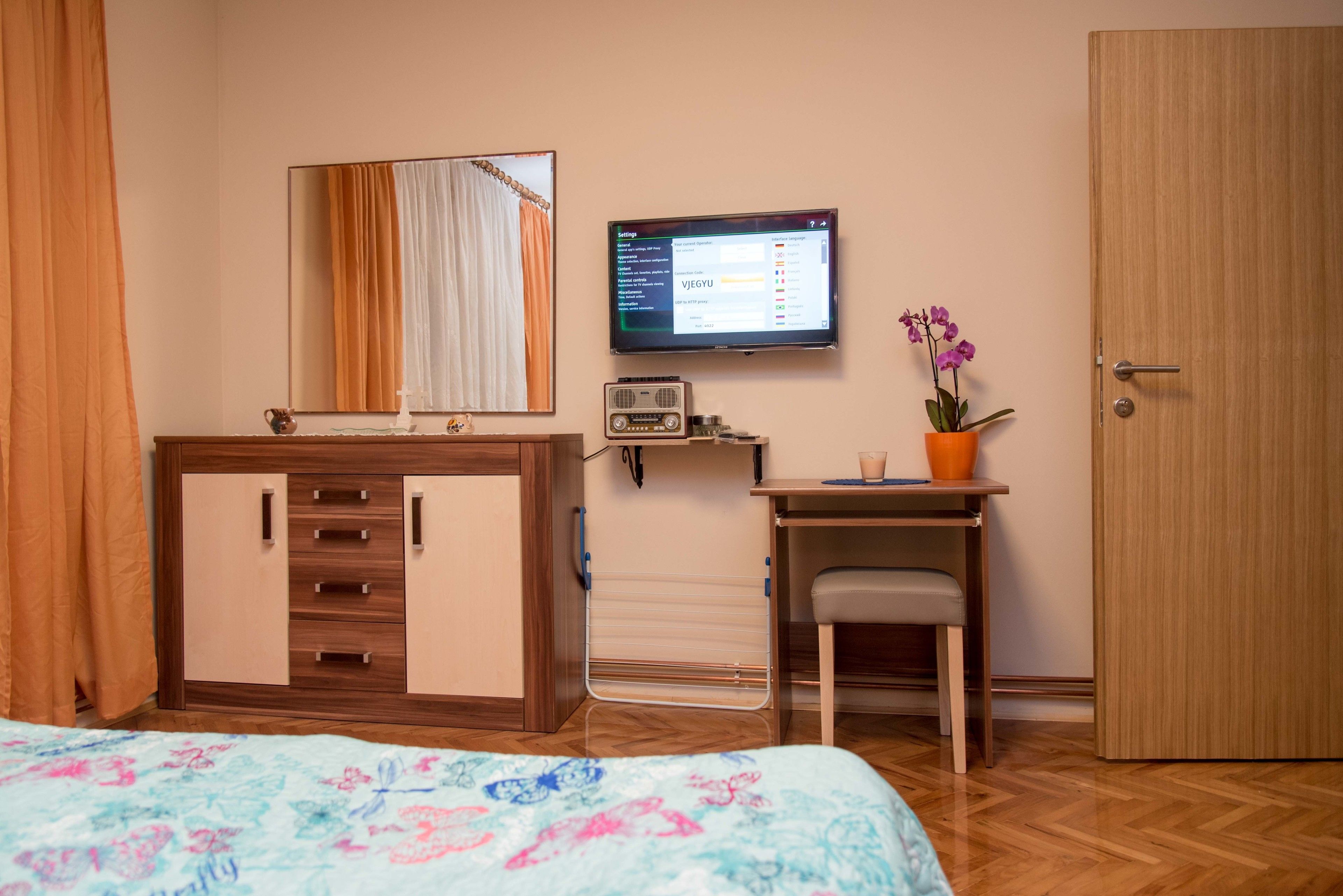 A bedroom with a bed, a wooden wardrobe, a desk with a chair, a television, and a potted plant.