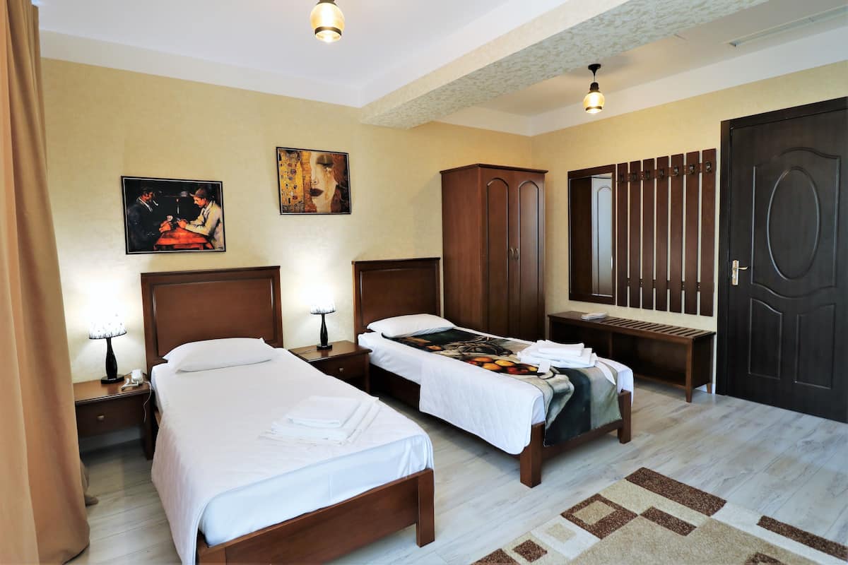 A hotel room with two single beds, a wooden wardrobe, a desk with a chair, a mirror, and two wall-mounted paintings.