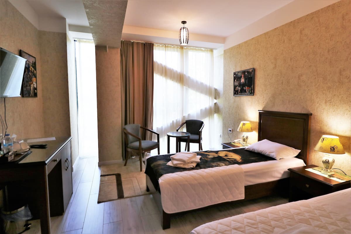 A hotel room with a bed, a desk, a chair, a small table, and a nightstand with a lamp.