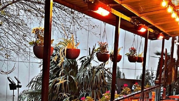 Un área de estar al aire libre con mesas redondas y sillas de mimbre, rodeada de macetas colgantes con diversas plantas.