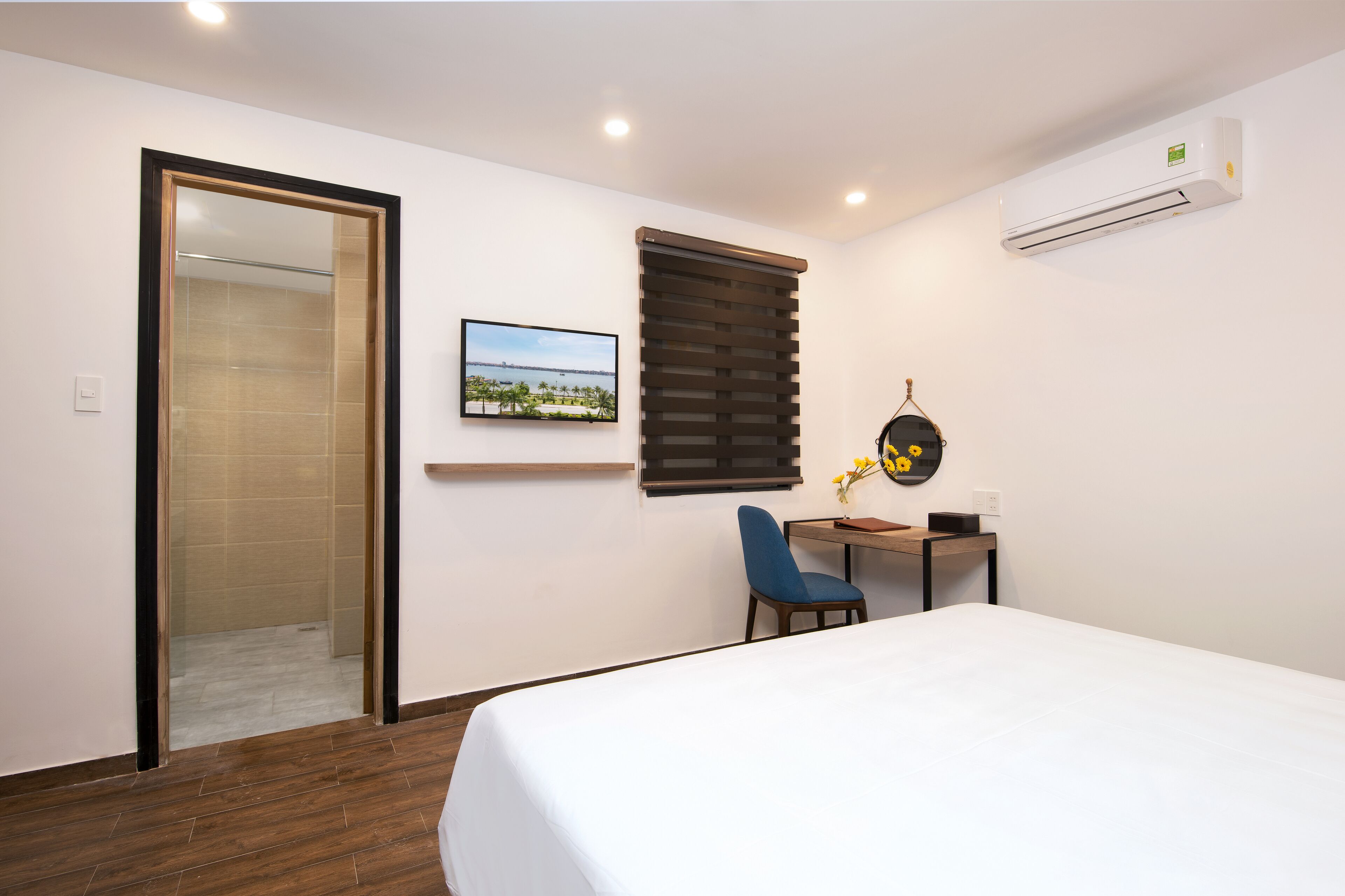 Habitación de hotel con cama, escritorio con silla, ventana con persianas, aire acondicionado y un cuadro en la pared.