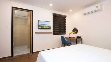 Habitación de hotel con cama, escritorio con silla, ventana con persianas, aire acondicionado y un cuadro en la pared.
