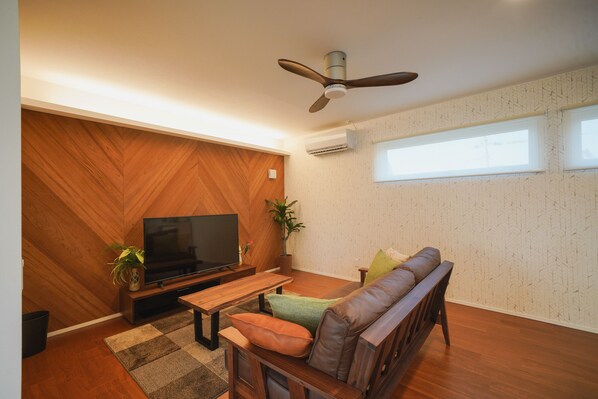 A modern living room with a wooden herringbone wall, a ceiling fan, a flat-screen TV, a brown leather sofa, and a wooden coffee table.
