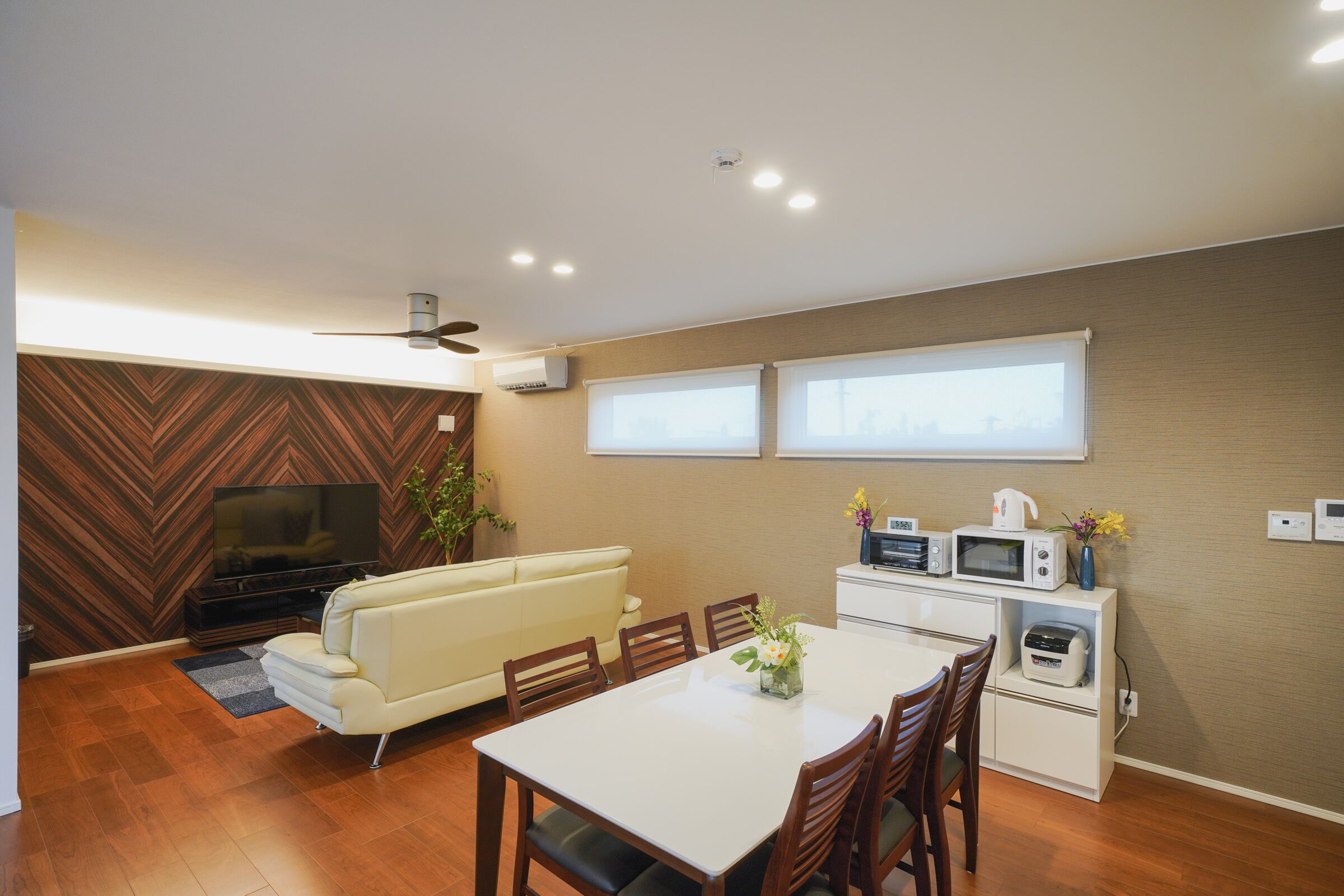A modern living room with a flat-screen TV, a sofa, a dining table with chairs, and a kitchen area with a microwave and toaster.