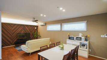 A modern living room with a flat-screen TV, a sofa, a dining table with chairs, and a kitchen area with a microwave and toaster.
