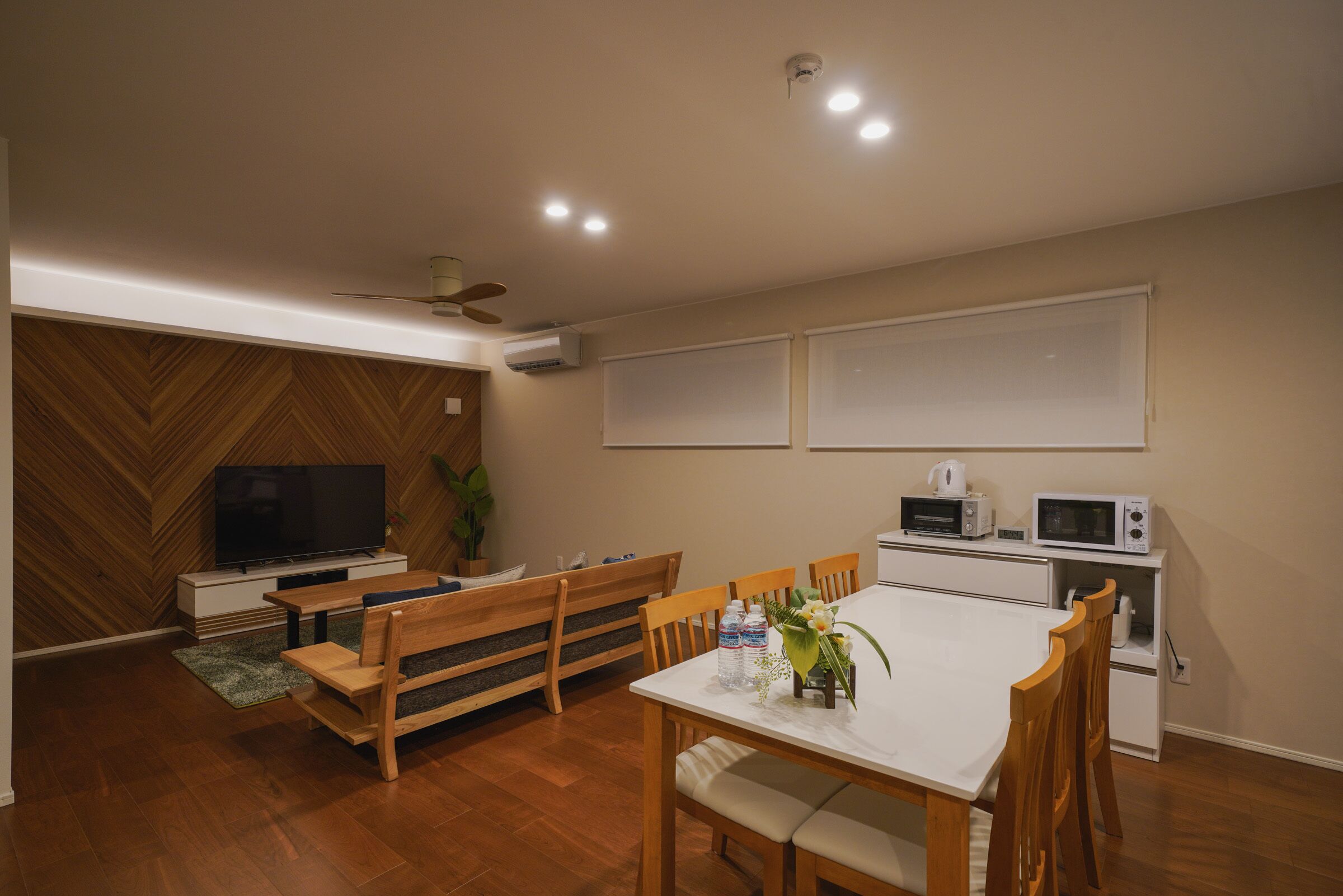 Una sala de estar moderna con una pared de madera en patrón de espiga, un televisor de pantalla plana, una mesa de comedor con sillas, un banco y una zona de cocina con microondas y hervidor.