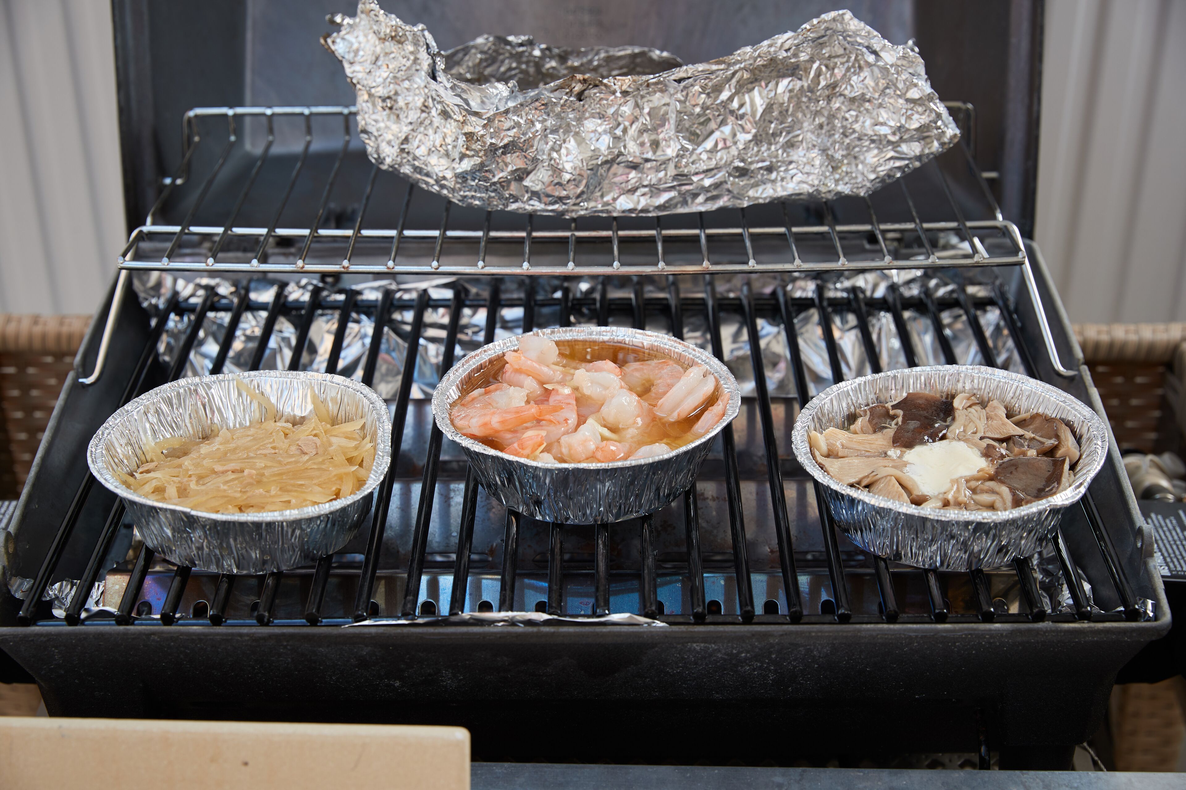 Three aluminum foil pans on a grill with food.