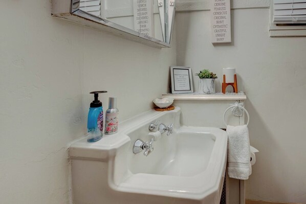 A bathroom with a white bathtub, a sink, and a mirror above. There are toiletries on the sink and a framed picture on the wall.