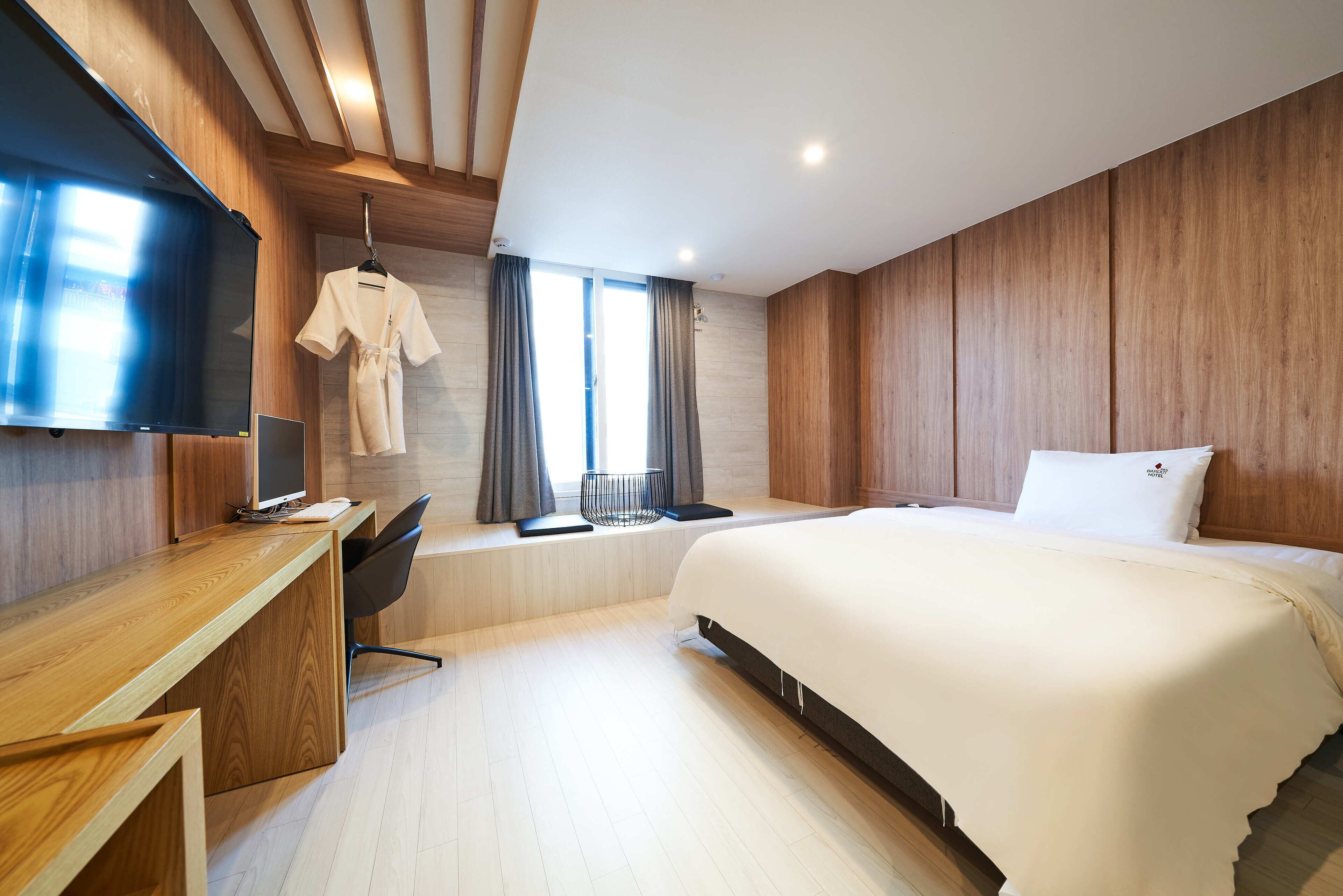 A modern hotel room with a wooden desk, a flat-screen TV, a bed with white bedding, and a large window with curtains.
