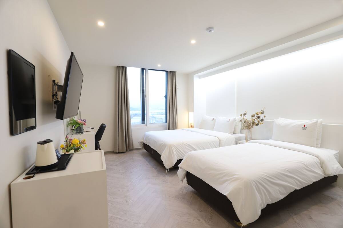 A hotel room with two beds, a desk with a coffee maker, a flat-screen TV, and a large window with curtains.