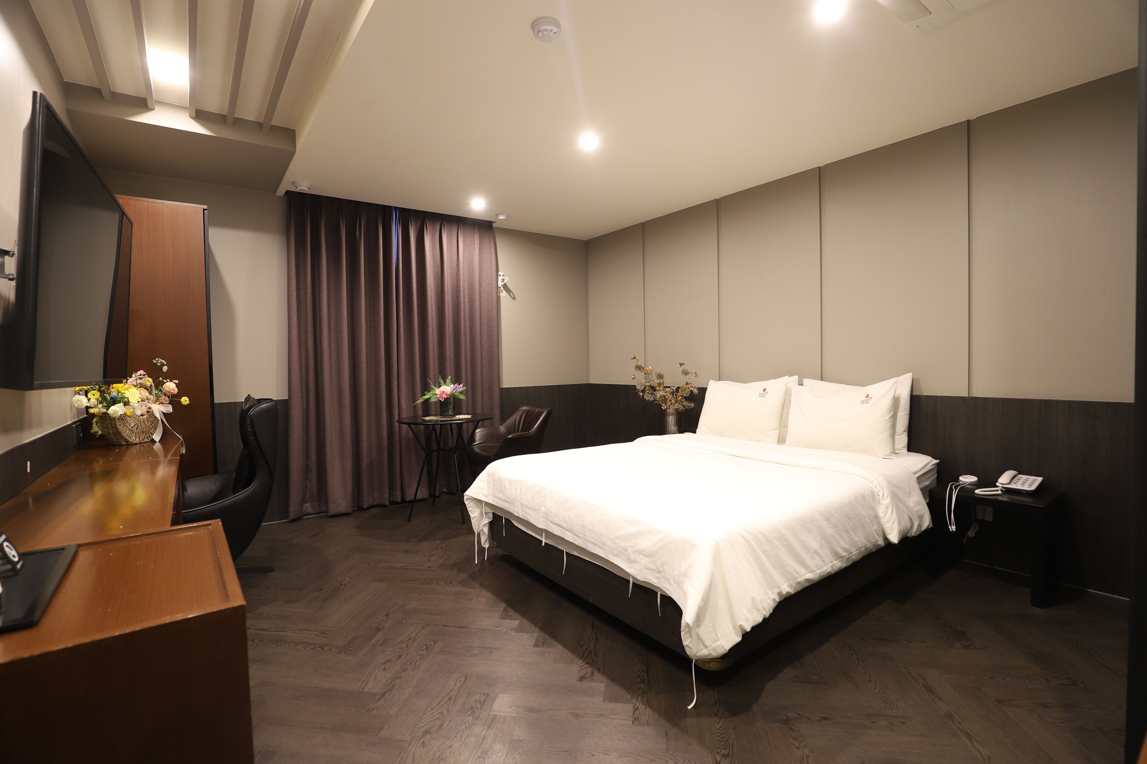 Quarto de hotel moderno com uma cama grande, uma escrivaninha, uma cadeira, uma mesinha com flores e uma mesa de cabeceira com um telefone.