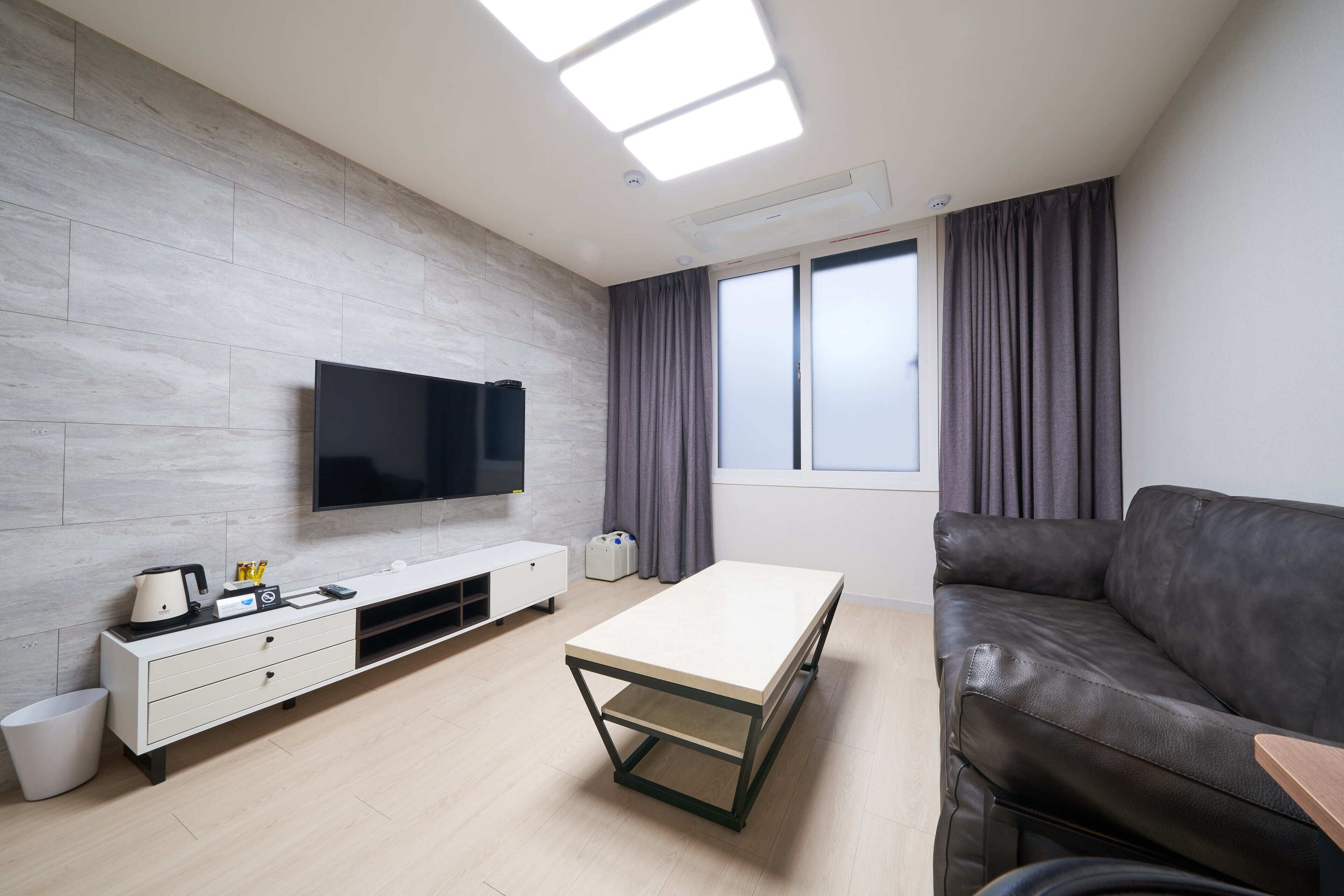 A modern living room with a flat-screen TV mounted on the wall, a white console table, a black leather sofa, and a large window with curtains.