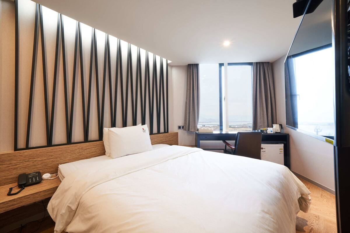 A hotel room with a large bed, a desk, a chair, a telephone, and a view of the city through the window.