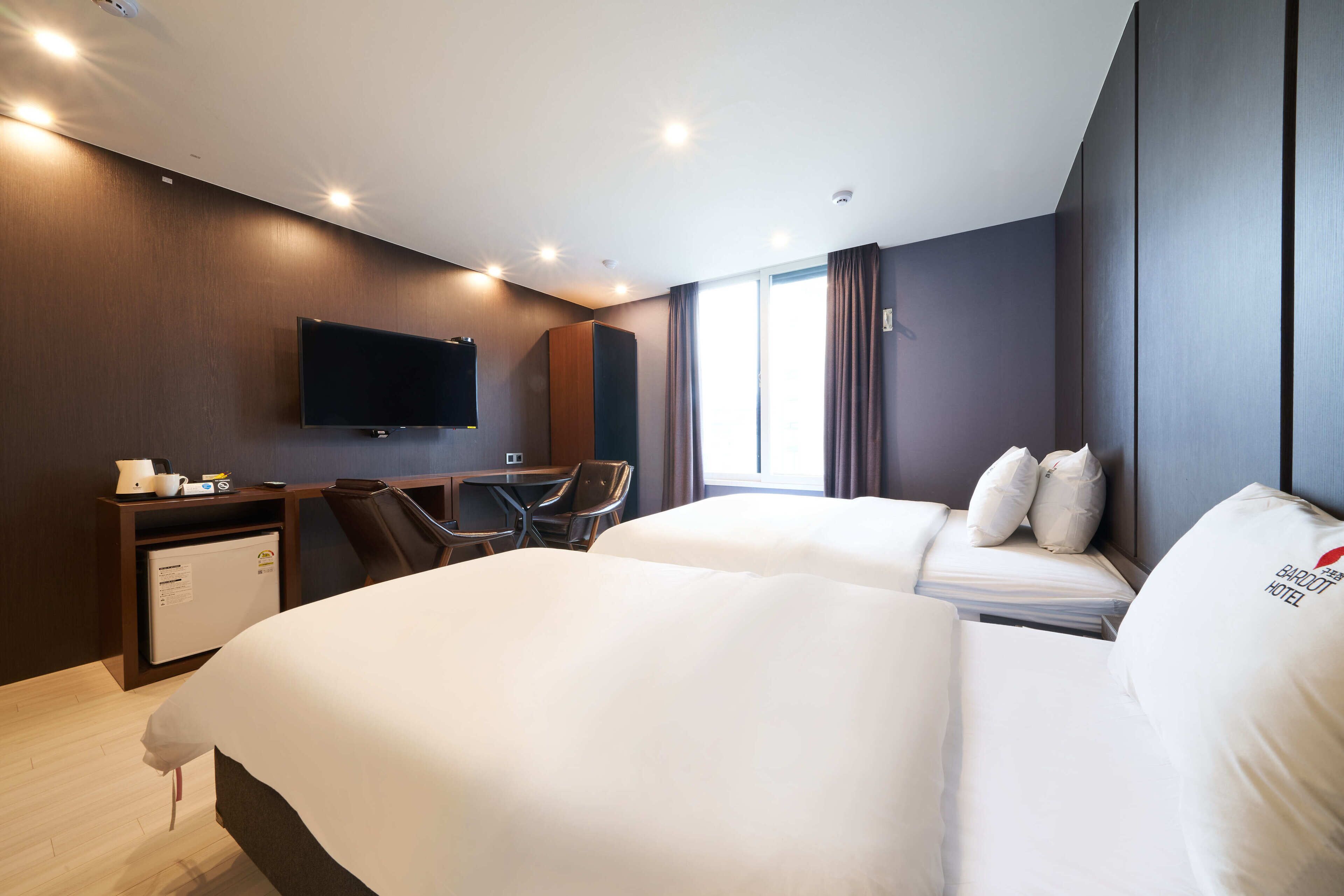 Una habitación de hotel con dos camas, un escritorio con silla, televisión y ventana con cortinas.