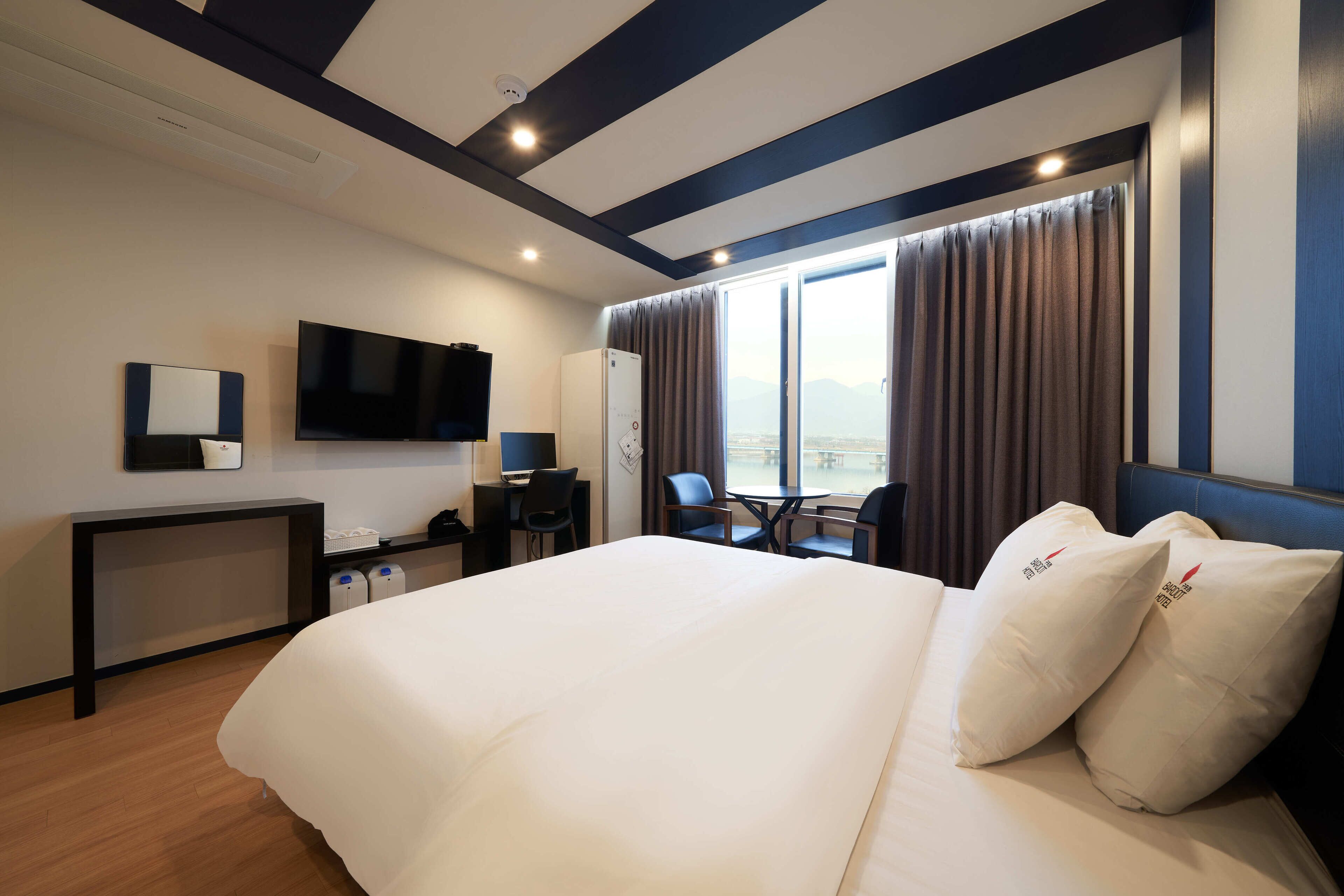 Quarto de hotel com uma cama grande, uma escrivaninha com computador, televisão e vista para as montanhas pela janela.