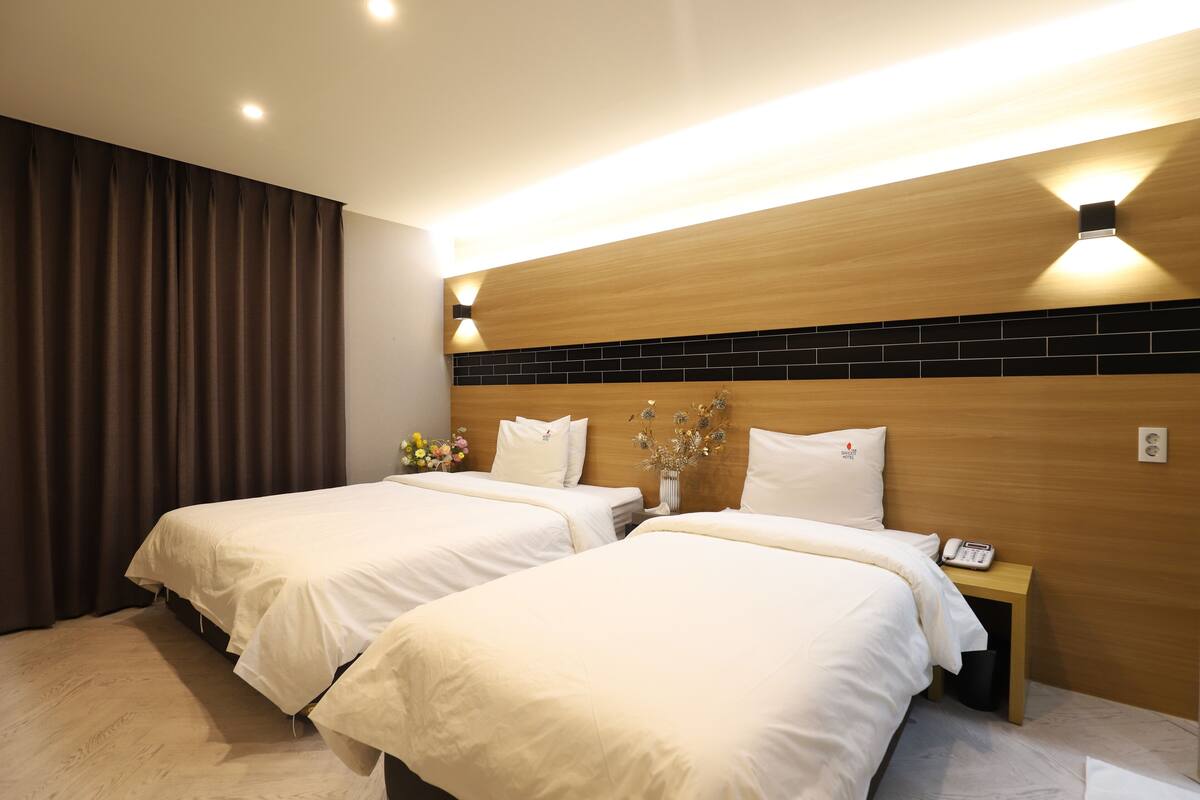 A hotel room with two beds, a nightstand with a phone, and a wall-mounted light fixture.