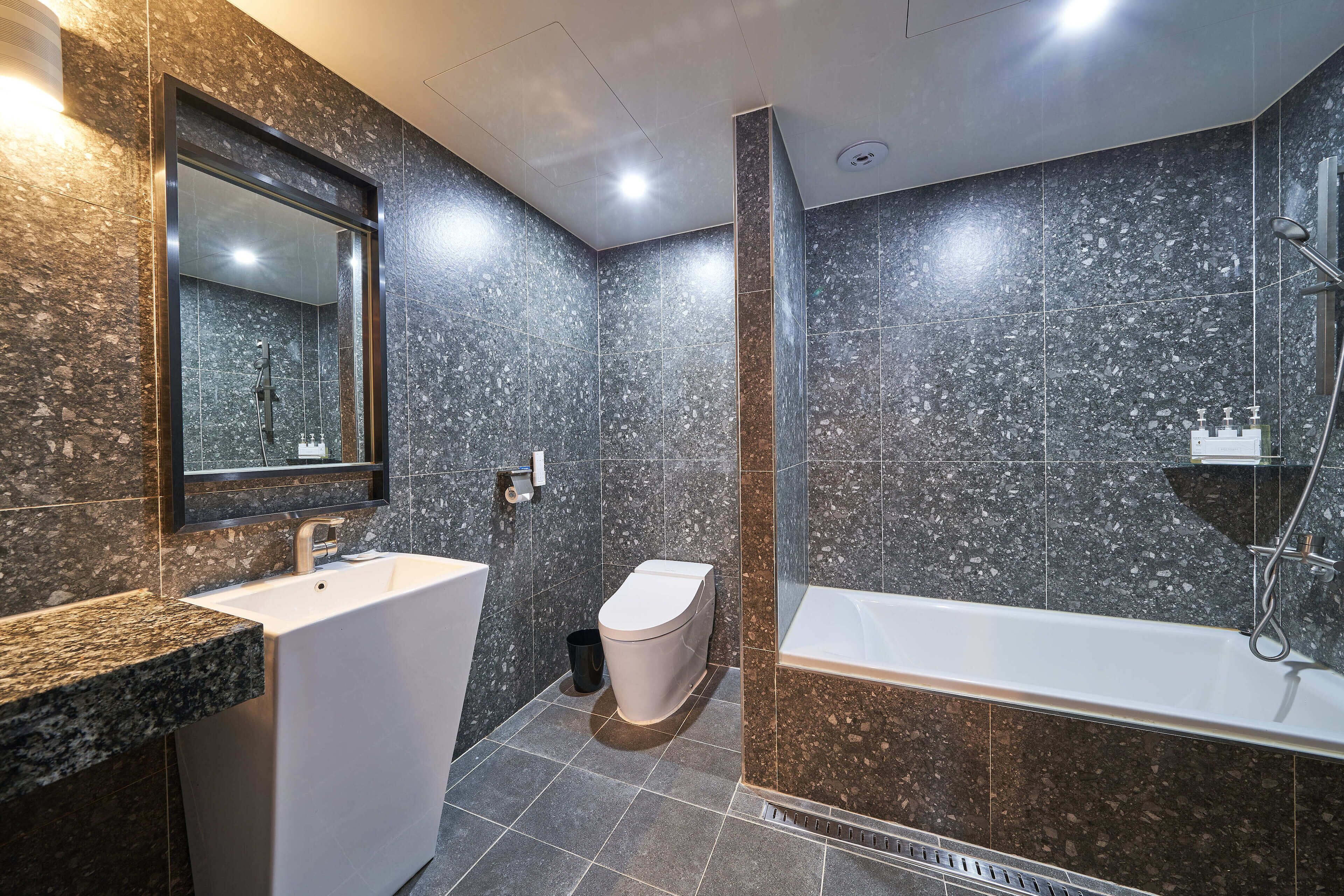 A modern bathroom with a white sink, toilet, and bathtub, all set against dark speckled tiles.