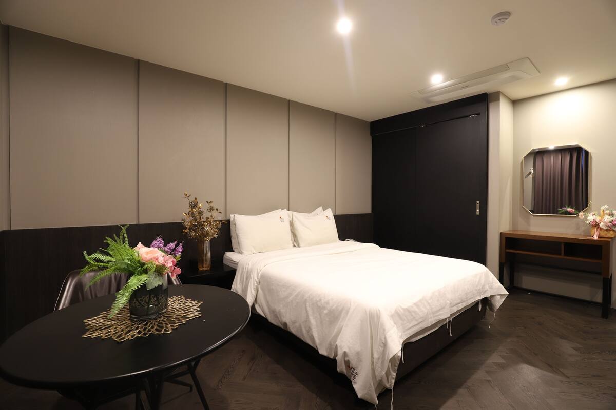 A modern hotel room with a large bed, a small round table with a vase of flowers, and a dark-colored wardrobe.