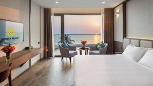 A hotel room with a bed, two armchairs, a small table with flowers, a TV on the wall, and a view of the sunset over the ocean. - Sunset Beach Resort & Spa- Beachfront Pirate Fire Show (Phu Quoc)