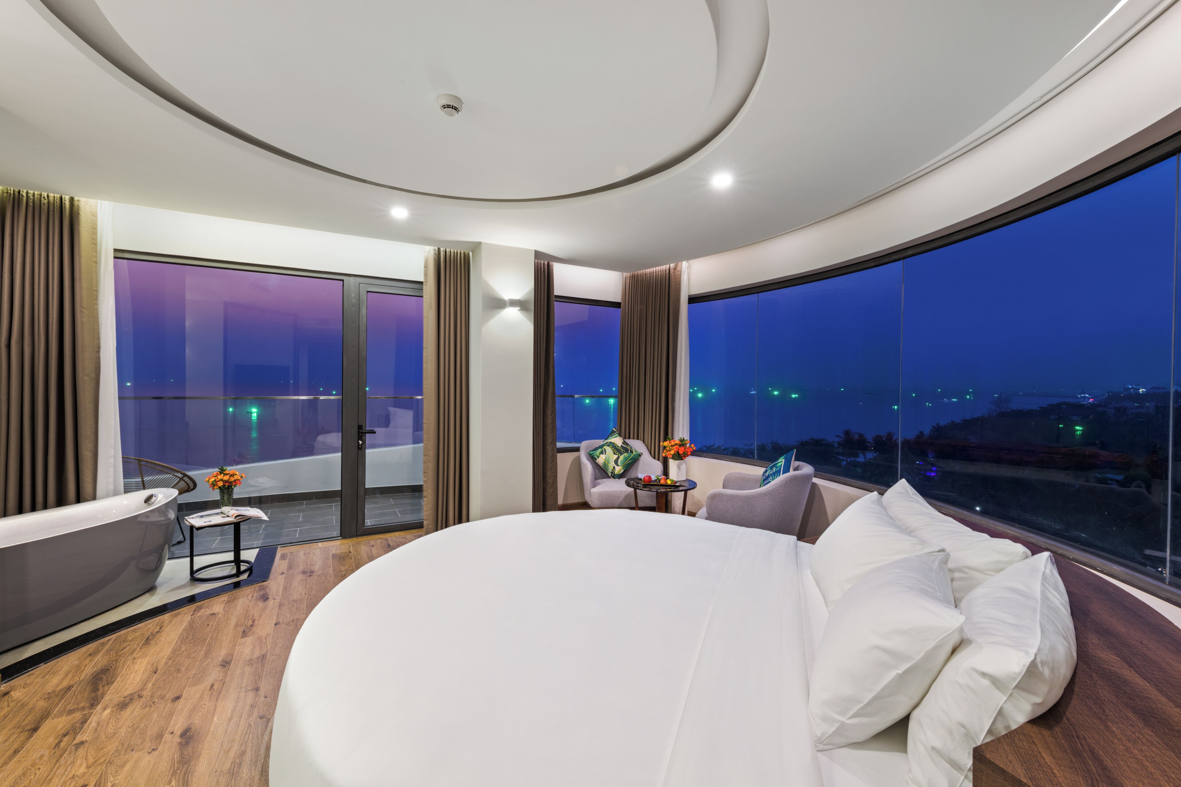 A modern hotel room with a large bed, a bathtub, a small table with flowers, and a view of the city at night.
