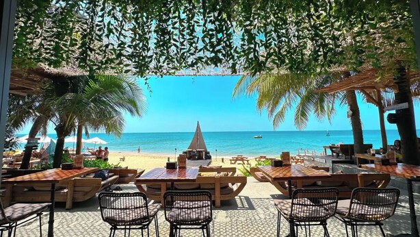 An outdoor seating area with tables and chairs, overlooking a beach with palm trees and a clear blue sky. - Sunset Beach Resort & Spa- Beachfront Pirate Fire Show (Phu Quoc)