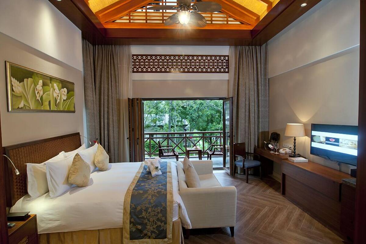 Habitación de hotel con cama, sofá, escritorio con televisor y balcón con mobiliario exterior.