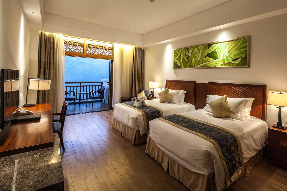 Habitación de hotel con dos camas, un escritorio, una silla, un balcón con vistas y un cuadro en la pared.