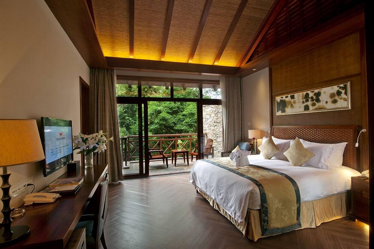 Habitación de hotel con cama, escritorio con computadora, televisor y balcón con vista a la vegetación.