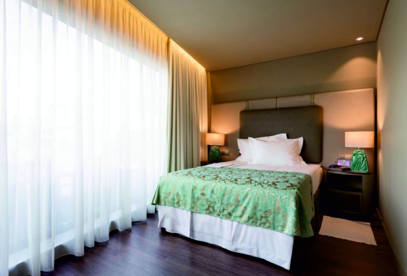 Ein Hotelzimmer mit einem großen Bett, einem Nachttisch mit Lampe und transparenten Vorhängen am Fenster.