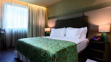 A hotel room with a bed, a desk, a chair, and a nightstand with a lamp.