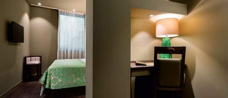 Una habitación de hotel con una cama, un escritorio con silla, una lámpara y un televisor en la pared.