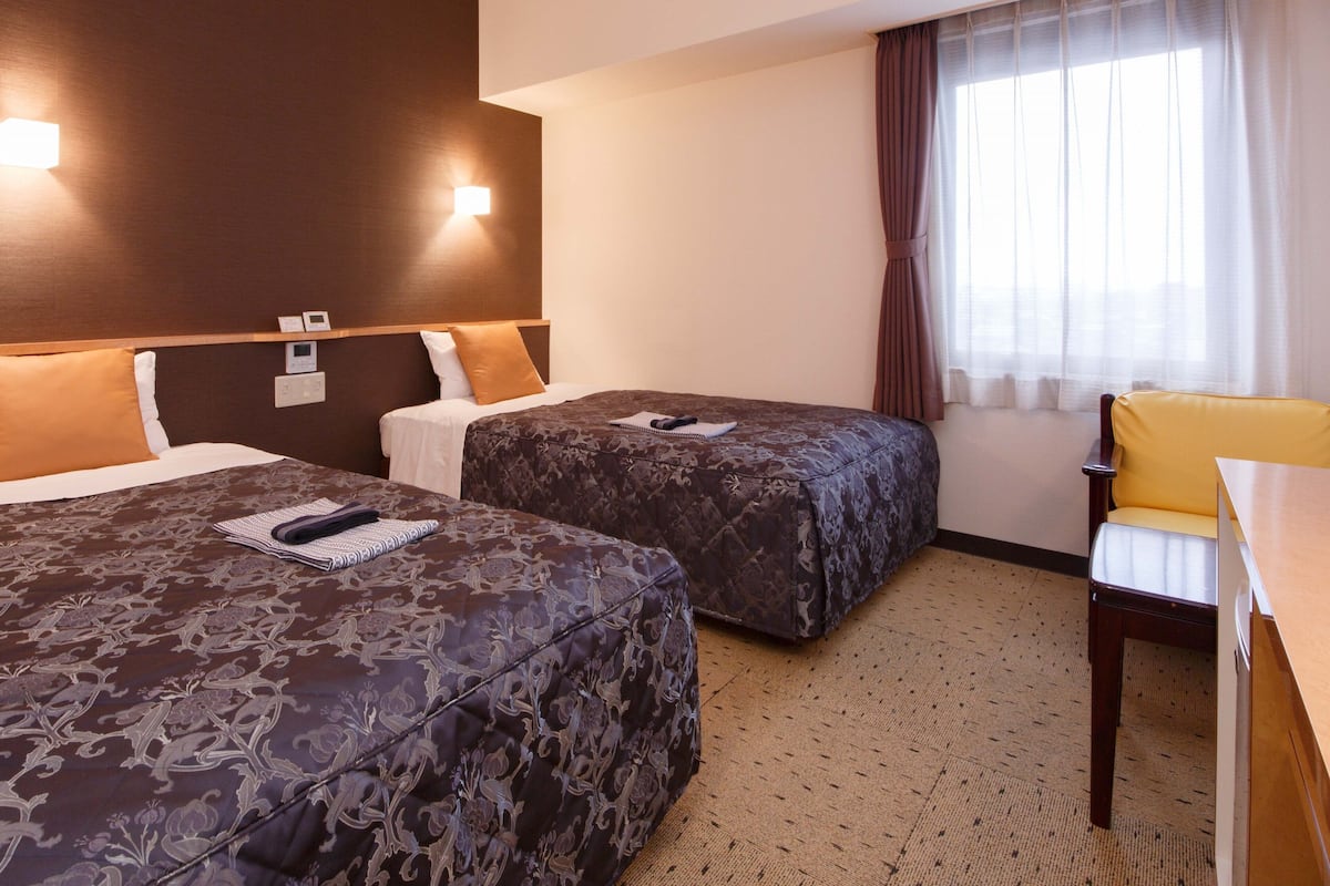Une chambre d’hôtel avec deux lits, un bureau, une chaise et une fenêtre avec des rideaux.