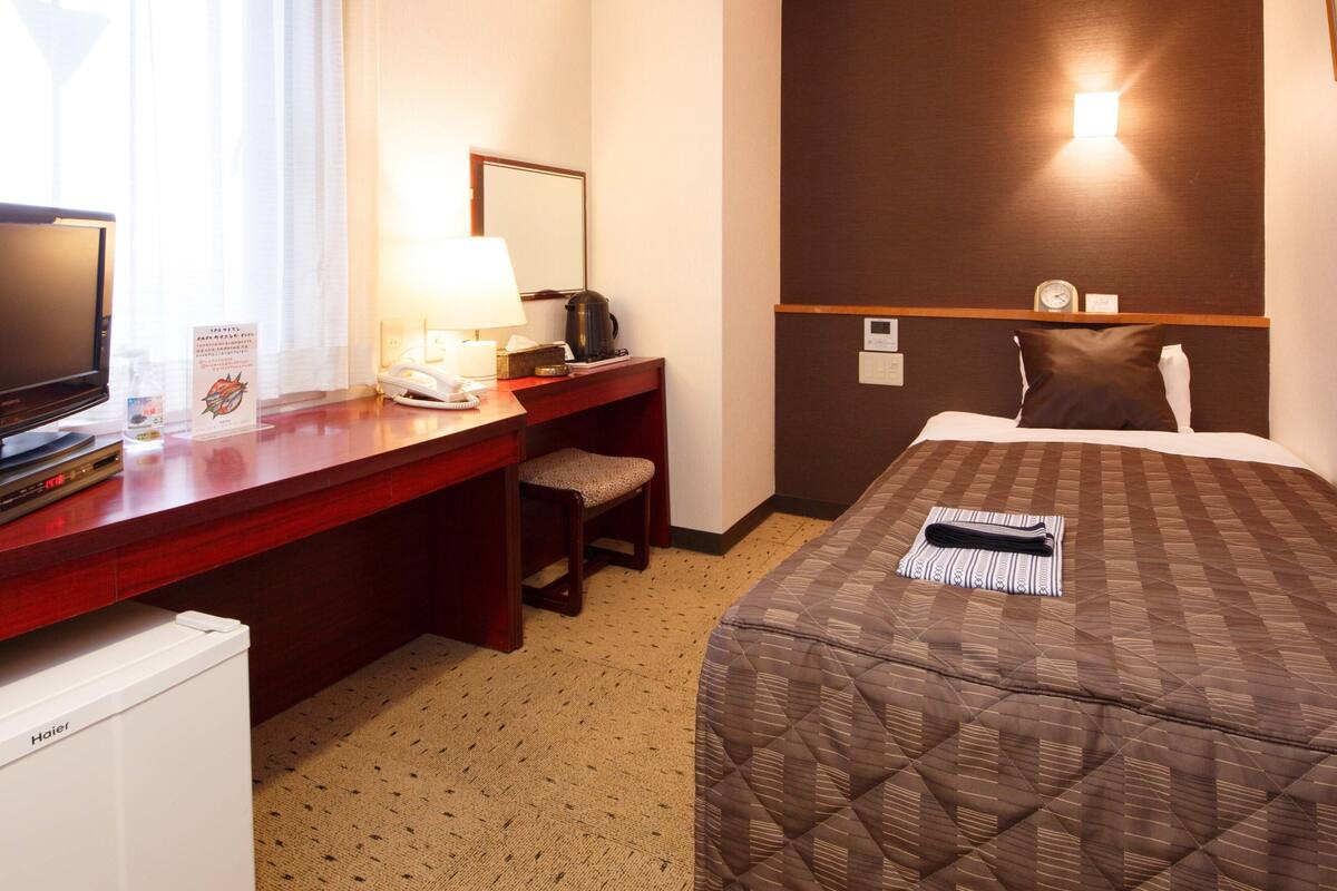 Een hotelkamer met een bed, een bureau met telefoon, een koffiezetapparaat, een televisie en een klok aan de muur.